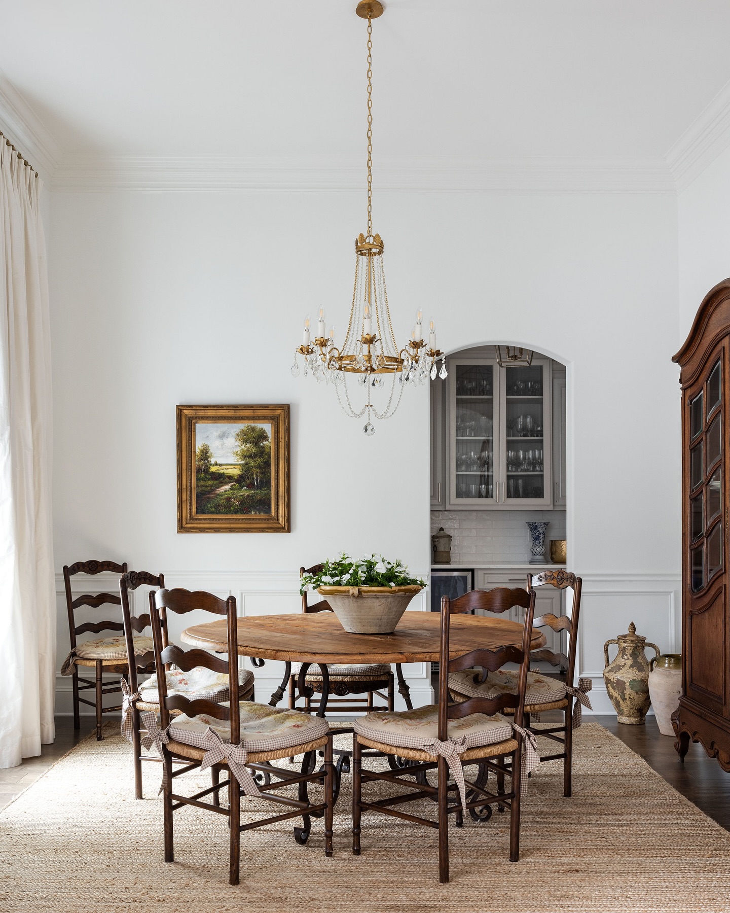 Dining room with a gilt crystal-swag chandelier over a round French farm table, ladder-back chairs and a carved walnut armoire