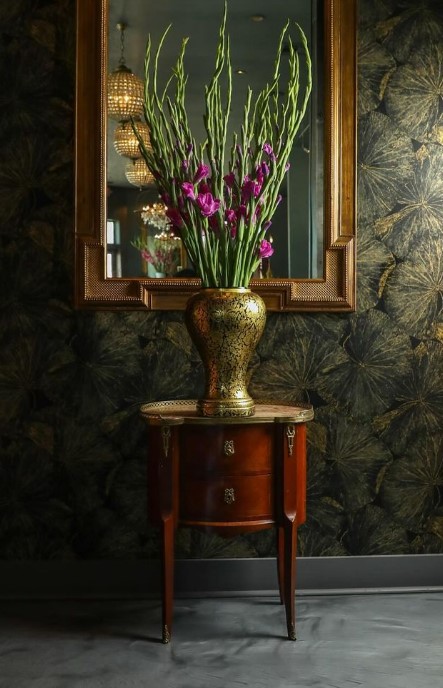 Entry vignette with a gilt-framed antique mirror above a mahogany demilune side table with brass mounts, holding a gilt-decorated vase of purple gladiolus against dark botanical wallpaper